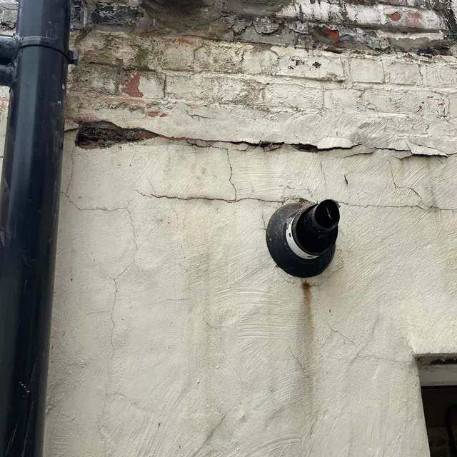 A boiler flue terminal coming through a rendered outside wall, beside a black soil pipe