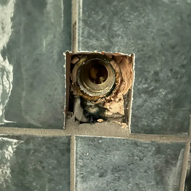 A brass shower valve seen through an opening cut in grey wall tiles